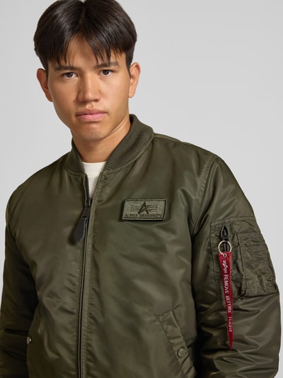 Männermodel trägt Jacke mit Reißverschlusstasche am Ärmel von Alpha Industries in Olivgrün mit Label-Patch, Nahaufnahme Oberkörper. Herren Jacke, Regular Fit, Kapuze mit Reißverschluss, gerippter Stehkragen, Reißverschluss, Eingrifftaschen mit Druckknopfverschluss, gerippte Abschlüsse, Rückenlänge 64 cm, Schulterbreite 54 cm, Brustweite 54 cm, Ärmellänge 72 cm.