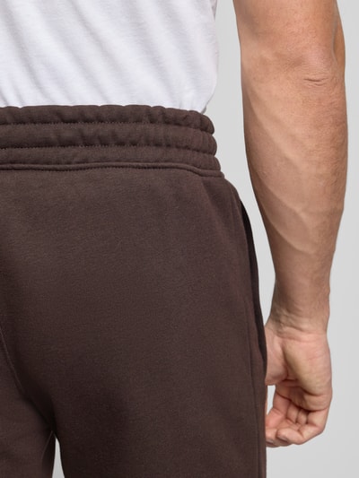 Männermodel draagt Sweatpants mit elastischem Bund van adidas Originals in donkerbruin katoenmix met steekzakken en labelstitching, detailaufnahme heup.
