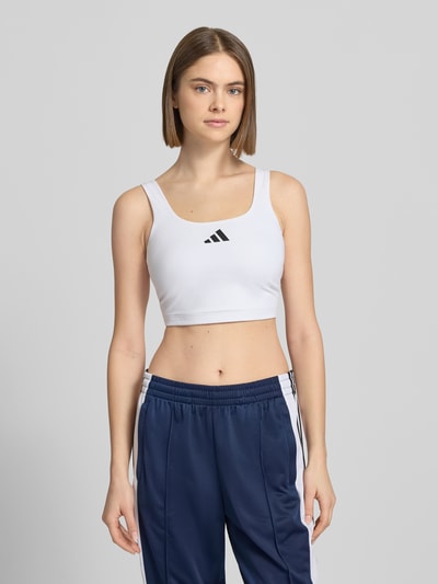 Vrouwenmodel draagt BH mit Label-Print van adidas Sportswear in wit met zwarte print, aansluitende fit, Materiaalmix met stretch, close-up bovenlichaam vooraanzicht.