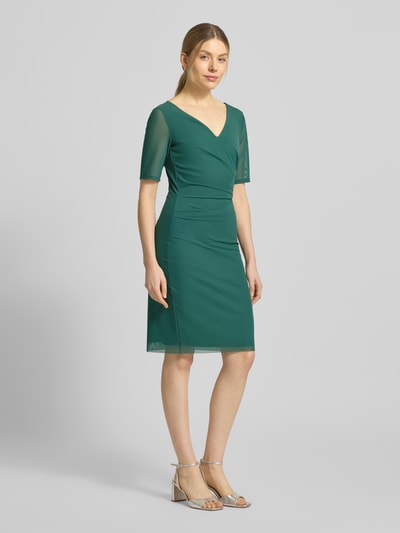 Frauenmodel trägt Regular Fit Midikleid in semitransparentem Design von Vera Mont in Dunkelgrün, V-Ausschnitt, kurzärmelig, Ganzkörperansicht von vorne. Elastischer Material-Mix, blickdichter Unterrock, Nahtreißverschluss, Rückenlänge 94 cm, Brustweite 37 cm, Ärmellänge 28 cm.