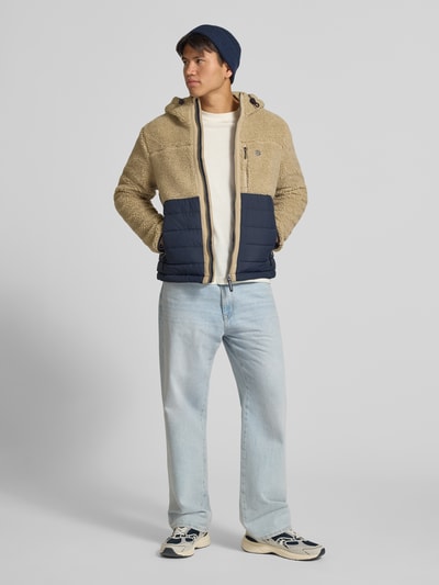 Mężczyzna nosi Regular Fit Jacke mit Teddyfell Modell 'Borg' od Superdry w beżowym i granatowym kolorze, krój regular fit, tekstura miękkiego teddy, kieszenie napoleonki i wpuszczane, kaptur ze sznurkiem, długość pleców w rozmiarze M: 64 cm, szerokość ramion w rozmiarze M: 45 cm, szerokość klatki piersiowej w rozmiarze M: 55 cm, długość rękawa w rozmiarze M: 68 cm, zdjęcie całej sylwetki od przodu.