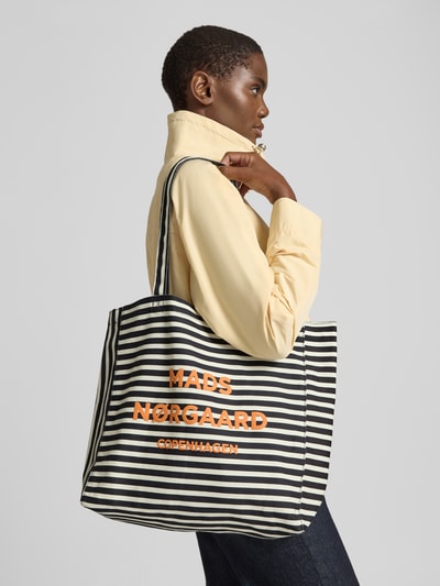 Frauenmodel trägt Shopper aus Baumwolle mit Logo Modell 'Boutiqno Athene' von MADS NØRGAARD, schwarz-weiß gestreift, mit orangefarbenem Logo-Print, sichtbare Tragehenkel, offene Struktur, Ganzkörperansicht von vorne. Breite 37 cm, Länge 35 cm, Tiefe 11 cm, Schulterriemen 61 cm.