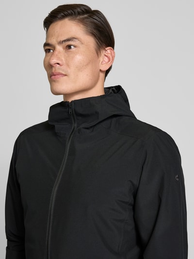 Männermodel trägt Regular Fit Jacke mit Kapuze von SCANDINAVIAN EDITION in Schwarz, glatte Textur, Zweiwege-Reißverschluss, Nahaufnahme Oberkörper.