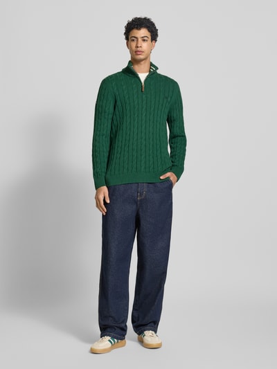 Männermodel trägt gebreide pullover uit puur katoen van Superdry met schipperskraag, kabelpatroon, labeldetail en ribboorden, heren, aansluitende pasvorm, ruglengte maat M 70 cm, mouwlengte maat M 71 cm, groen, donkerblauwe broek, sneakers, geheel van voren.