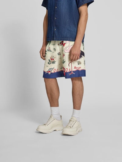 Männermodel trägt Seidenshorts aus reiner Seide von Dsquared2 in Creme mit bunten floralen und Tiermotiven, lockere Passform, glänzende Optik, Nahaufnahme Unterkörper.