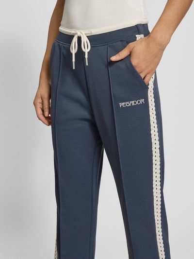 Kobieta nosi spodnie typu track pants Pegador Trackpants mit Spitzenbesatz Modell 'TIHANY LACE', mieszanka bawełny, krój regular fit, obszycie koronką, elastyczny pas, wyhaftowane logo, długość wewnętrzna nogawki w rozmiarze S: 77 cm, zbliżenie na górną część nogawek z przodu.
