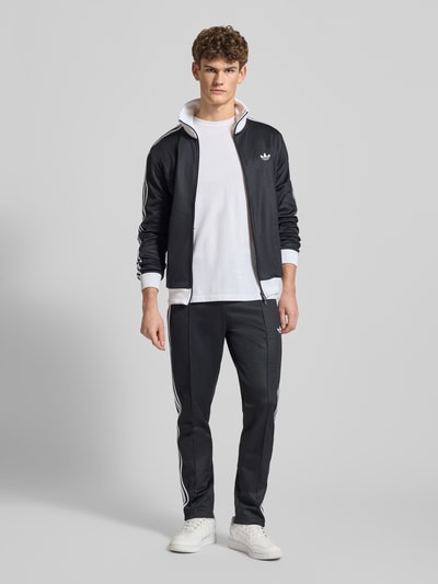 Männermodel trägt Regular Fit Trainingsjacke mit Stehkragen Modell 'Classic' von adidas Originals im Baumwoll-Mix mit Logo-Stitching, Zweiwege-Reißverschluss, elastischen Abschlüssen, ganz in dunklen Farben, Ganzkörperansicht von vorne.