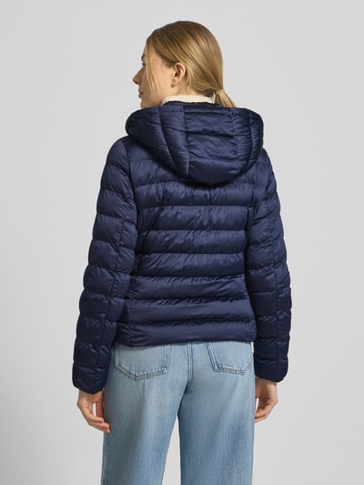 Frauenmodel trägt taillierte Steppjacke mit Zweiwege-Reißverschluss Modell 'ANYA' von Beaumont Amsterdam in Dunkelblau, Ganzkörperansicht Rückansicht. Damen Steppjacke mit Kapuze, Tunnelzug, kurze Rückenlänge, glatte Textur, leicht glänzend.