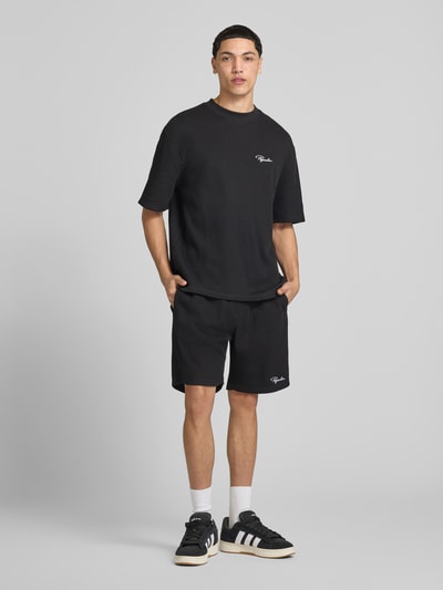 Männermodel trägt Shorts mit Logo-Stitching und elastischem Bund von Pegador aus Reine merzerisierte Baumwolle, Loose-Fit, Feinripp, mit französischen Eingrifftaschen und Gesäßtasche in Schwarz, Ganzkörperansicht von vorne.
