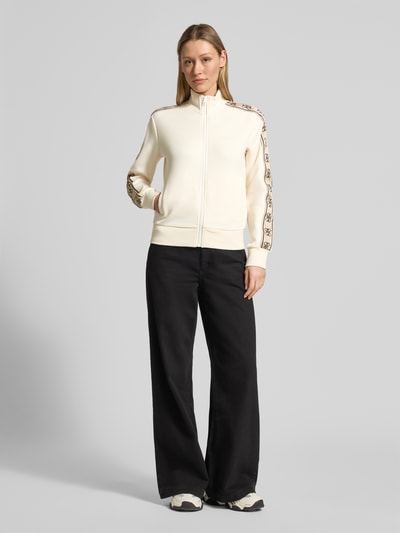 Frauenmodel trägt Sweatjacke mit Stehkragen und Eingrifftaschen Modell 'BRITNEY' von Guess Activewear, cremefarben, Regular Fit, mit Zierstreifen und Logo, Baumwoll-Mix, Reißverschluss, Ganzkörperansicht von vorne.