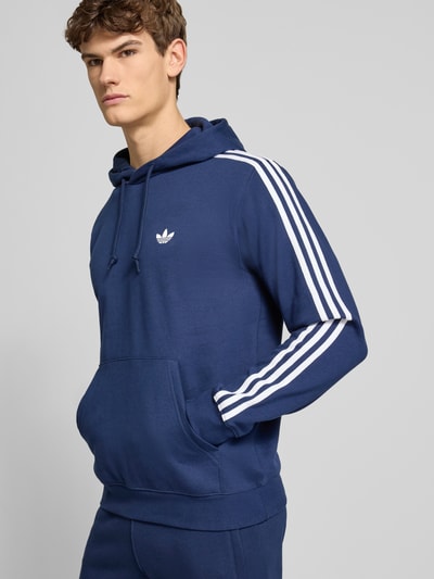 Mężczyznamodel nosi Hoodie mit Kapuze von adidas Originals z bawełnianego materiału, luźny krój, kaptur z troczkiem, granatowy z białymi paskami, kieszeń kangurka, ściągacze, długość tyłu w rozmiarze M 70 cm, długość rękawa 67 cm, widok całego ciała z przodu.