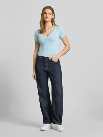 Frauenmodel trägt Cropped T-Shirt mit V-Ausschnitt und Knopfleiste von Guess Jeans im Material-Mix mit Viskose-Anteil, hellblau, Logo, Rückenlänge bei Größe S 42 cm, Ganzkörperansicht von vorne.