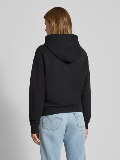 Modelka nosi Hoodie mit Label- und Motiv-Print Modell 'DAISY FLAG' von Tommy Jeans, czarny, luźny krój, bawełniany materiał z dodatkiem stretchu, jednolity kolor, kaptur ze sznurkiem, opuszczone ramiona, kieszeń kangurka, ściągacze. Widok całej sylwetki z tyłu.