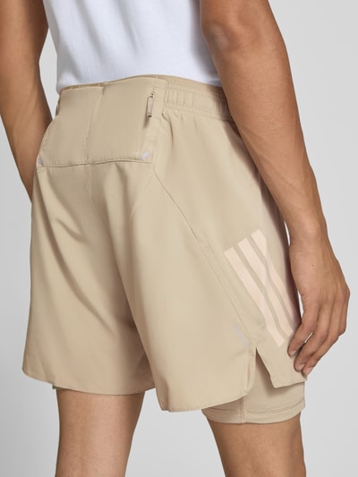 Männermodel trägt beigefarbene Regular Fit Sportshorts mit Logo-Print Modell 'ADI365/// 2i1 M' von adidas aus der adidas Training Kollektion, glatte Textur, dezenter Glanz, Ganzkörperansicht Rückansicht.