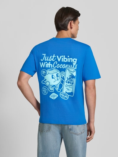 Männermodel trägt Regular Fit T-Shirt aus reiner Baumwolle Modell 'VIBING COCO' von REVIEW, Rückseite mit Print, blaue Farbe, Ganzkörperansicht Rückansicht. Herren T-Shirt von REVIEW x ANSON´S, reine Baumwolle, Rückenlänge bei Größe M: 68 cm.