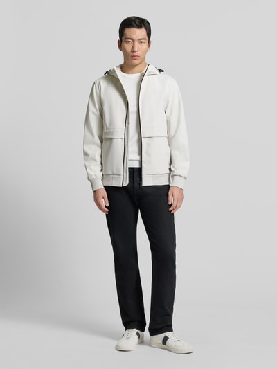 Męski model nosi Regular Fit Funktionsjacke mit Kapuze od Tom Tailor Denim, jednolity kolor, krój regular fit, wodoodporna, oddychająca, izolująca ciepło, stójka z kapturem i troczkiem, długie rękawy z elastycznym mankietem, zamek błyskawiczny, label stitching, elastyczny dół, pełny widok z przodu.