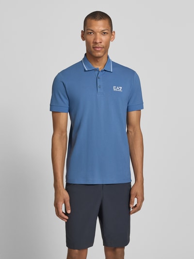 Męski model nosi Regular Fit Poloshirt mit Logo-Print od EA7 Emporio Armani, bawełniany elastyczny krój regular fit, kolor niebieski z białym nadrukiem logo, krótkie rękawy, polokołnierzyk, elastyczne ściągacze rękawów, długość pleców w rozmiarze M: 69 cm, widok całej sylwetki z przodu.