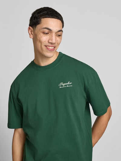 Männermodel trägt Oversized Fit T-Shirt mit Label-Detail Modell 'Morcote' von Pegador aus Reine Baumwolle, Grün, Halbarm, Nahaufnahme Oberkörper.