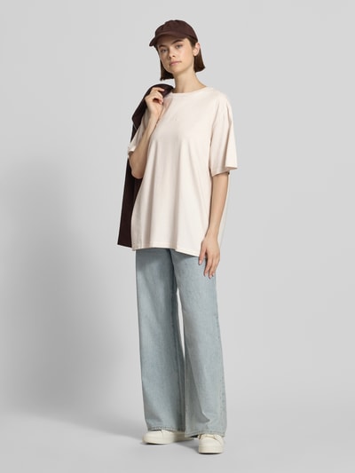 Frauenmodel trägt Oversized T-Shirt mit Label-Stitching von OH APRIL in Beige aus reiner Baumwolle, kombiniert mit Jeans, Ganzkörperansicht von vorne.