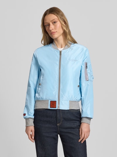 Frauenmodel trägt Bomberjacke mit Label-Patch und Reißverschlusstasche am Ärmel von Original Bombers, hellblau mit grauen elastischen Abschlüssen, seitliche Eingrifftaschen mit Druckknopfverschluss, Stehkragen, Logo-Stitching, Rückenlänge bei Größe S: 53 cm, Schulterbreite: 37 cm, Brustweite: 48 cm, Ärmellänge: 63 cm, Lockerer-Schnitt, Ganzkörperansicht von vorne.