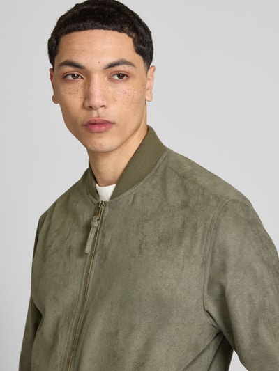 Męski model nosi bomberkę in Leder-Optik mit Stehkragen von Jack & Jones, kolor oliwkowy, krój regular fit, zamek błyskawiczny, wpuszczane kieszenie, długość tyłu w rozmiarze M: 64 cm, szerokość ramion w rozmiarze M: 45 cm, szerokość klatki piersiowej w rozmiarze M: 52 cm, długość rękawów w rozmiarze M: 67 cm, półzbliżenie przód.