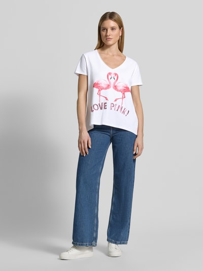 Kobiece model prezentuje damski t-shirt z mieszanki bawełny i elastanu, krój regular fit, model 'Flamingo Love Me' od miss goodlife z dekoltem w serek i nadrukiem z motywem, długość tyłu w rozmiarze S: 66 cm, widok całej sylwetki od przodu.