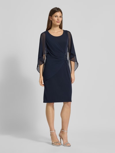 Frauenmodel trägt knielanges Cocktailkleid mit Rundhalsausschnitt von Vera Mont in Dunkelblau, Wickel-Optik, Fledermausärmel aus Chiffon, Strassstein-Applikation, körpernahe Passform, elastischer Material-Mix, Ganzkörperansicht von vorne.