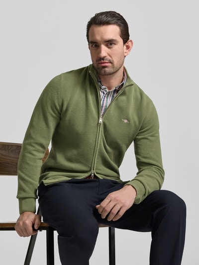 Männermodel trägt Cardigan mit Zweiwege-Reißverschluss und Label-Stitching von Gant in Grün aus reiner Baumwolle, Slim-Fit, Stehkragen, Ganzkörperansicht von vorne.