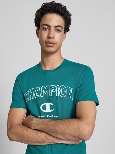 Männermodel trägt T-Shirt mit Label-Print und Rundhalsausschnitt von CHAMPION in Grün, reine Baumwolle, Regular Fit, Nahaufnahme Oberkörper.