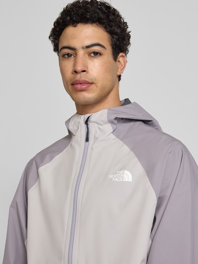 Männermodel trägt Regenjacke mit Logo von The North Face in hellgrau mit Kapuze, Reißverschluss, Regular Fit, Nahaufnahme Oberkörper. Rückenlänge bei Größe M: 70 cm, Schulterbreite: 50 cm, Brustweite: 62 cm, Ärmellänge: 76 cm.