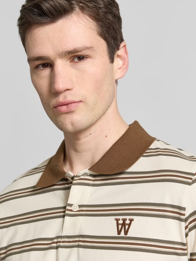 Męski model nosi Slim Fit Poloshirt mit Label-Stitching Modell 'Colton' von Wood Wood z bawełny, krój slim fit, wyhaftowane logo, listwa guzikowa, kremowo-brązowe pasy, Nahaufnahme Oberkörper.