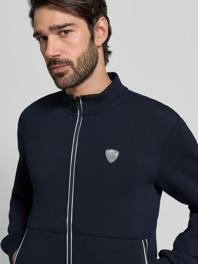 Männermodel trägt Sweatjacke mit Reißverschluss von EA7 Emporio Armani in Dunkelblau mit Label-Detail, Regular-Fit, Nahaufnahme Oberkörper. Rückenlänge bei Größe M: 70 cm, Ärmellänge: 66 cm, Stehkragen, Gerippte-Abschlüsse, Eingrifftaschen.
