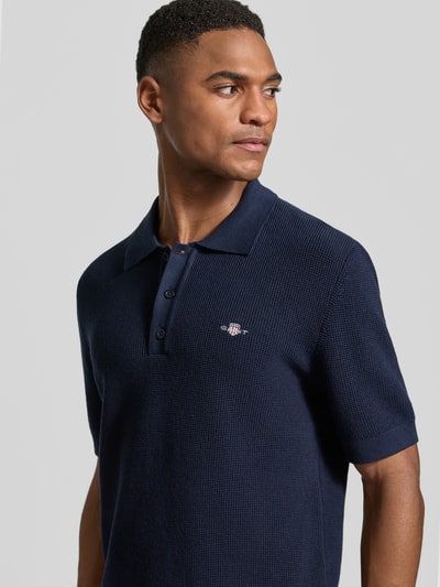 Męski model nosi Regular Fit Poloshirt mit Label-Stitching od Gant z bawełny, kołnierzyk polo, krój regular fit, wyhaftowane logo, długość tyłu w rozmiarze M: 68 cm, zbliżenie na górną część ciała z przodu
