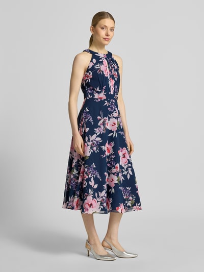 Frauenmodel trägt Midikleid mit floralem Allover-Print von Christian Berg Cocktail in Dunkelblau mit Rosa und weißen Blüten, Taillierte Passform, Rundhalsausschnitt, Gelegte Falten, Ärmelloses-Design, sichtbarer Nahtreißverschluss, Ganzkörperansicht von vorne.