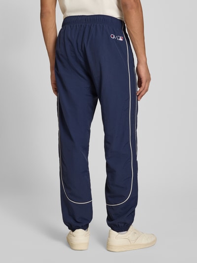 Männermodel trägt Relaxed Fit Tracksuit mit Logo-Patch von Champion in Dunkelblau mit weißen Paspeln, elastische Abschlüsse, Ganzkörperansicht Rückansicht. Herren Tracksuit, Reißverschluss, Stehkragen, Bundweite 39 cm, Rückenlänge 68 cm, Schulterbreite 42 cm, Ärmellänge 49 cm, Innenbeinlänge 72 cm.