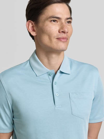 Mężczyznamodel nosi Regular Fit Poloshirt mit Brusttasche und Label-Stitching von bugatti w kolorze jasnoniebieskim, kołnierzyk polo, listwa guzikowa, wyhaftowane logo, brustasza, długość tyłu w rozmiarze M 70 cm, koszulka polo o kroju regular fit; zbliżenie na górną część ciała z przodu.