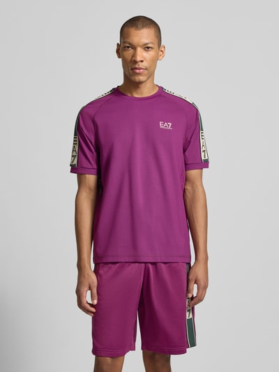 Męski model trägt T-shirt aus Viskose-Mix mit Logo von EA7 Emporio Armani, krój regular fit, okrągły dekolt, mieszanka wiskozy z dodatkiem streczu, logo, długość tyłu w rozmiarze M 70 cm, kolor fioletowy z zielonymi wstawkami, Ganzkörperansicht von vorne.
