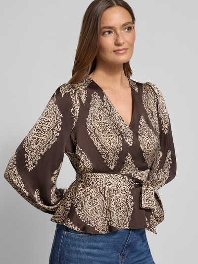 Modelka nosi bluzkę 'Regular Fit Bluse mit Allover-Paisley-Print Modell 'Rista' von Neo Noir, krój regular fit, długi rękaw, dekolt w serek, kopertowy styl, taliowany, wzór paisley na całej powierzchni, długość tyłu 54 cm, długość rękawów 63 cm, ciemnobrązowe i beżowe odcienie; zbliżenie na górną część ciała z przodu.