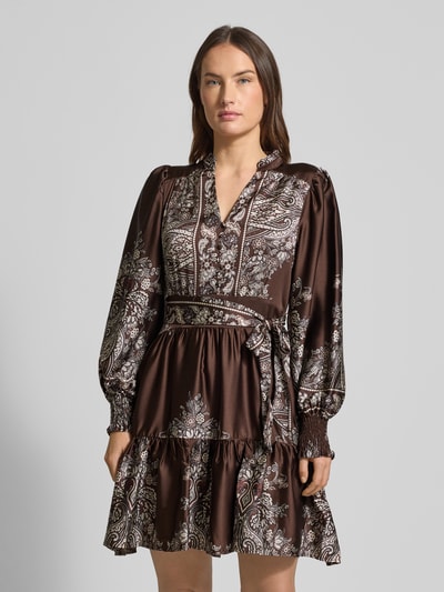 Kobiece modelka nosi sukienkę do kolan z dekoltem w serek model 'MALFIDA' od Neo Noir, taliowany krój, pasek w talii, krótka listwa guzikowa, długość tyłu 92 cm, szerokość klatki piersiowej 43 cm, długość rękawów 64 cm, wzór brązowo-biały, połyskująca tekstura, widok od przodu, ujęcie całej sylwetki.