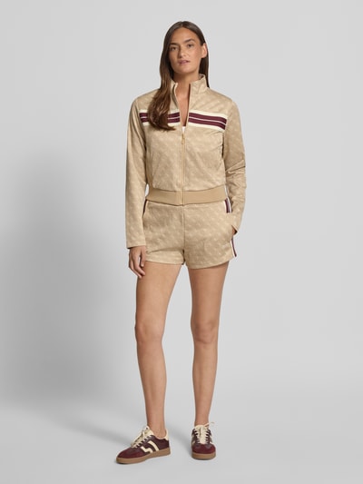 Frauenmodel trägt Shorts mit Allover-Logo-Print Modell 'Jacquard' von Guess Activewear, ausgestellte Passform, elastischer Bund, beige mit Logo-Muster, Material-Mix mit Baumwoll-Anteil, Eingrifftaschen, Ganzkörperansicht von vorne.