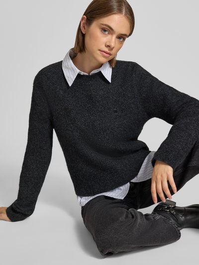 Vrouwenmodel draagt gebreide pullover met labeldetail Model 'Curtis' van Carhartt Work In Progress, materiaalmix met wol, getailleerde pasvorm, geribde ronde hals en ribboorden, ruglengte maat S: 55 cm, mouwlengte maat S: 60 cm, donkere tinten, zachte textuur, ganzebovenaanzicht van voren