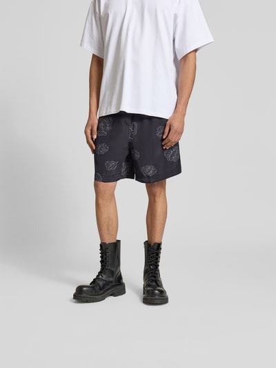 Männermodel trägt Shorts aus reiner Viskose von Officine Générale in Schwarz mit Muster, elastischem Bund und Tunnelzug, Ganzkörperansicht von vorne.