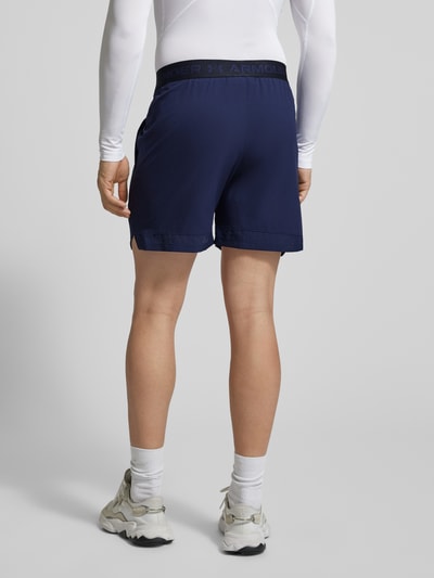Mannenmodel draagt Straight fit short met logoprint model 'Vanish' van Under Armour in donkerblauw, elastische band, steekzakken, bandwijdte maat M 38 cm, binnenbeenlengte maat M 15 cm, geheel van achteren.