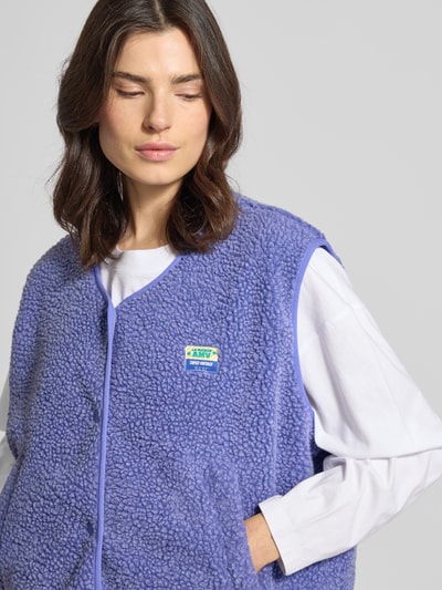 Frauenmodel trägt Weste mit Rundhalsausschnitt Modell 'HOKTOWN' von American Vintage in Lila, Teddyfell, Druckknopfleiste, Label-Detail, Eingrifftaschen, Lockerer-Schnitt, Nahaufnahme Oberkörper.