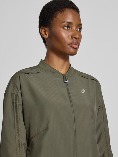 Modelka nosi kurtkę treningową z elastycznej mieszanki materiałów Model 'NAGINO' od ASICS TIGER, khaki, odblaskowe detale, zamek błyskawiczny, luźny krój, nahaufnahme górnej części ciała.