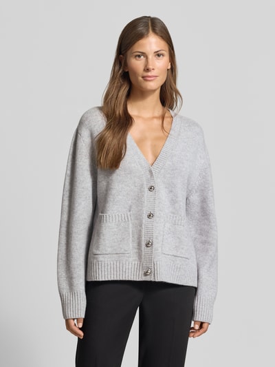 Frauenmodel trägt Strickjacke aus Woll-Mix mit Kaschmir-Anteil von MYON in Hellgrau, lockere Passform, Knopfleiste, aufgesetzte Taschen, gerippte Abschlüsse, überschnittene Schultern, Ganzkörperansicht von vorne.