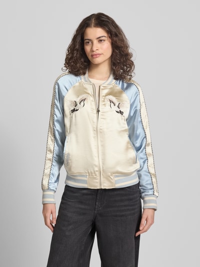 Modelka kobieca nosi Bomber Jacke mit Motiv- und Logo-Stitching od Superdry, satynowa tkanina, kremowe i błękitne kolory, prosty krój, krótka stójka, zamek, boczne kieszenie, elastyczne wykończenia, długość tyłu w rozmiarze S: 58 cm, szerokość ramion w rozmiarze S: 45 cm, szerokość klatki piersiowej w rozmiarze S: 53 cm, długość rękawa w rozmiarze S: 64 cm, widok całej sylwetki od przodu