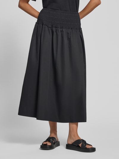 Frauenmodel trägt Midirock mit Smok-Detail von Comma aus schwarzem Baumwoll-Mix in A-Linie, elastischer Bund, Nahaufnahme Unterkörper.