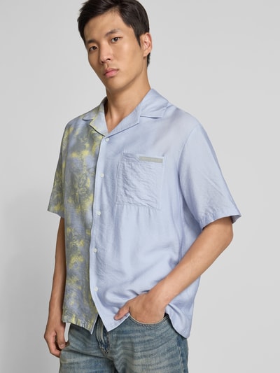 Männermodel trägt Relaxed Fit Freizeithemd aus Viskose-Mix von MAISON KITSUNÉ, hellblau mit Brusttasche, Durchgehende Knopfleiste, Kurzärmelig, Rückenlänge 73 cm, Schulterbreite 55 cm, Ärmellänge 28 cm, Kontraste in Gelb auf einer Seite, Ganzkörperansicht von vorne.