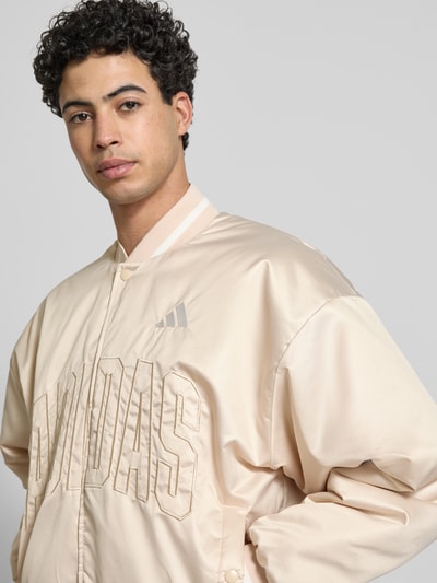 Männermodel trägt Regular Fit Bomberjacke mit Stehkragen von adidas Sportswear, beige, Logo-Stitching, Nahaufnahme Oberkörper. Rückenlänge 69 cm, Schulterbreite 64 cm, Brustweite 67 cm, Ärmellänge 61 cm, Druckknopfverschluss, elastische Abschlüsse.